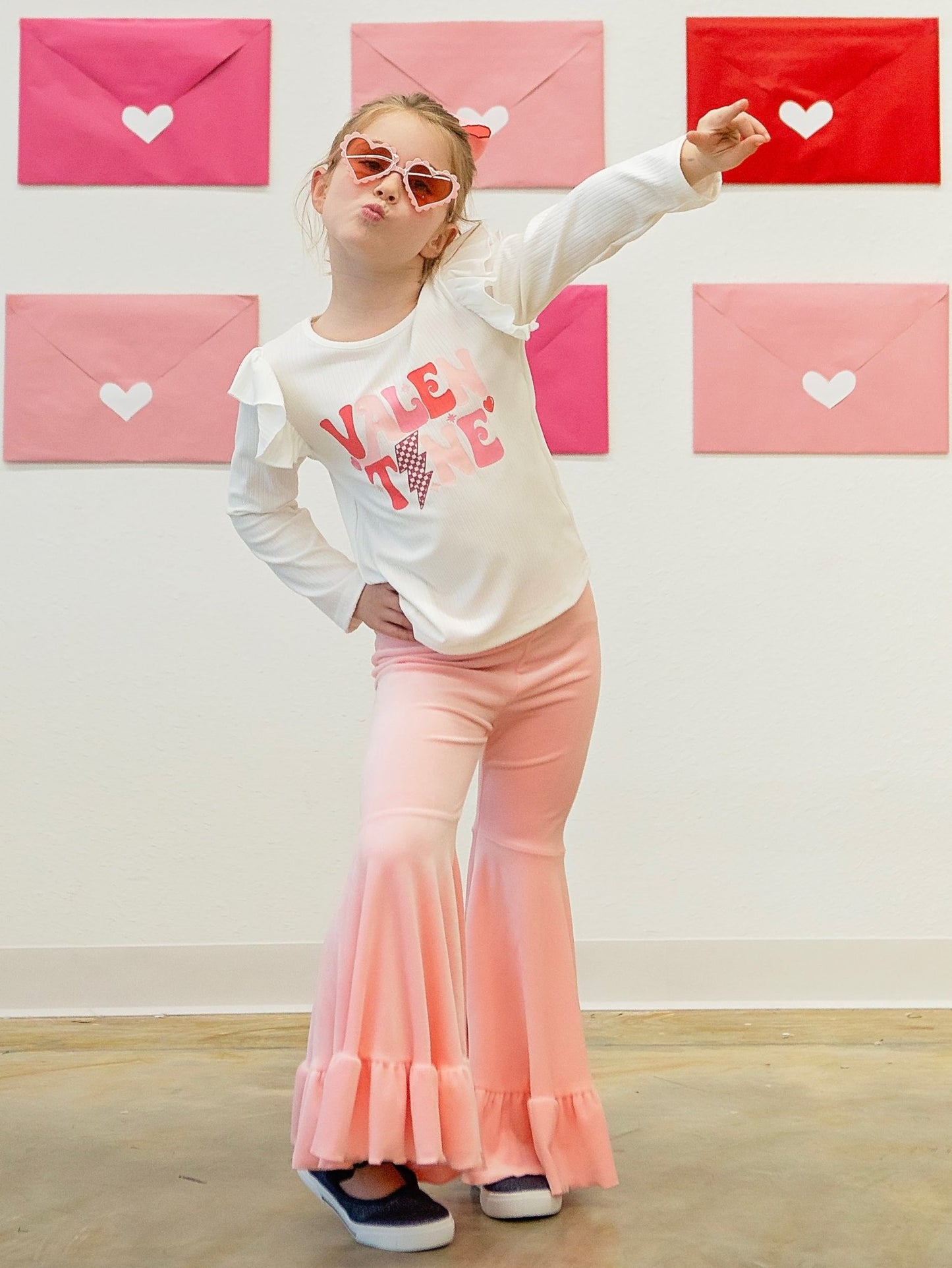 Model wearing a pink bell bottom set with a white top that has the text 'I love you' and a pair of sunglasses. The background displays Valentine's Day themed decorations with pink and red colors.