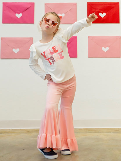 Model wearing a pink bell bottom set with a white top that has the text 'I love you' and a pair of sunglasses. The background displays Valentine's Day themed decorations with pink and red colors.