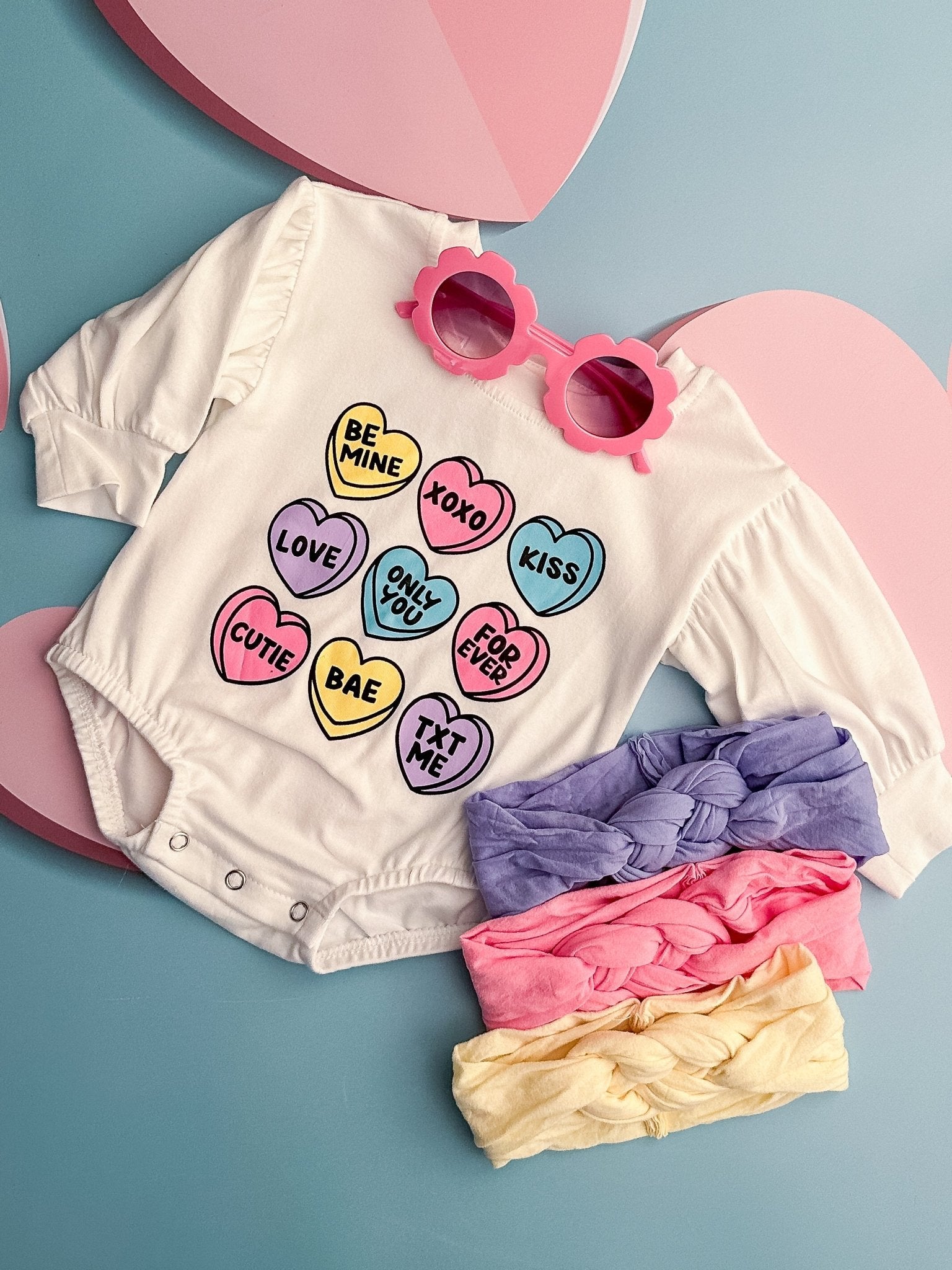 Valentine Sweetheart Infant Onesie displayed with heart-shaped decorations and accompanied by pink sunglasses and multi-colored bow ties.