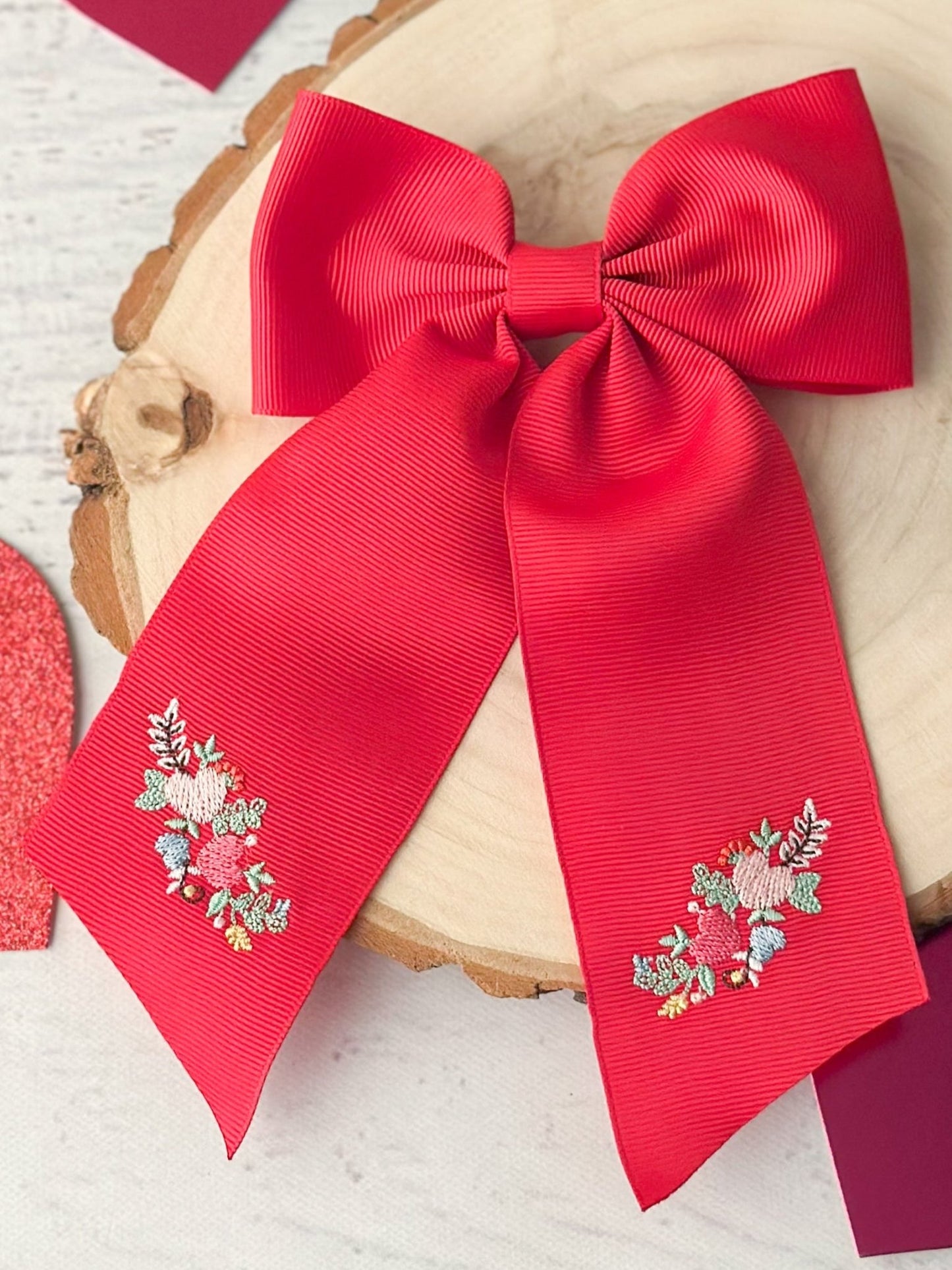 A red long tail hair bow with embroidered details on the ribbon, displayed on a wooden stand.