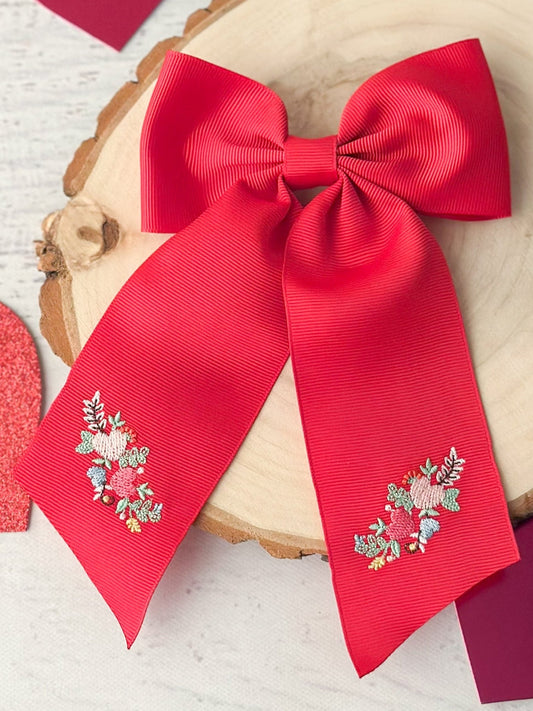 A red long tail hair bow with embroidered details on the ribbon, displayed on a wooden stand.
