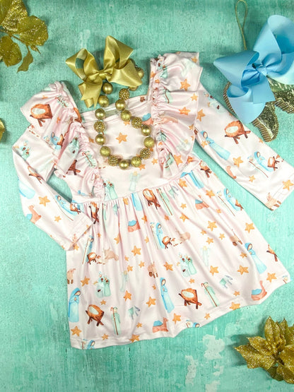 A light pink long-sleeve toddler dress with a gathered waist and ruffled shoulders, decorated with a nativity-themed print featuring stars, angels, Mary, Joseph, baby Jesus in a manger, and animals. The dress is laid flat on a teal background, accessorized with a large gold bow, a chunky gold beaded necklace, and a large blue bow nearby. Gold glitter poinsettia decorations are placed in the corners.
