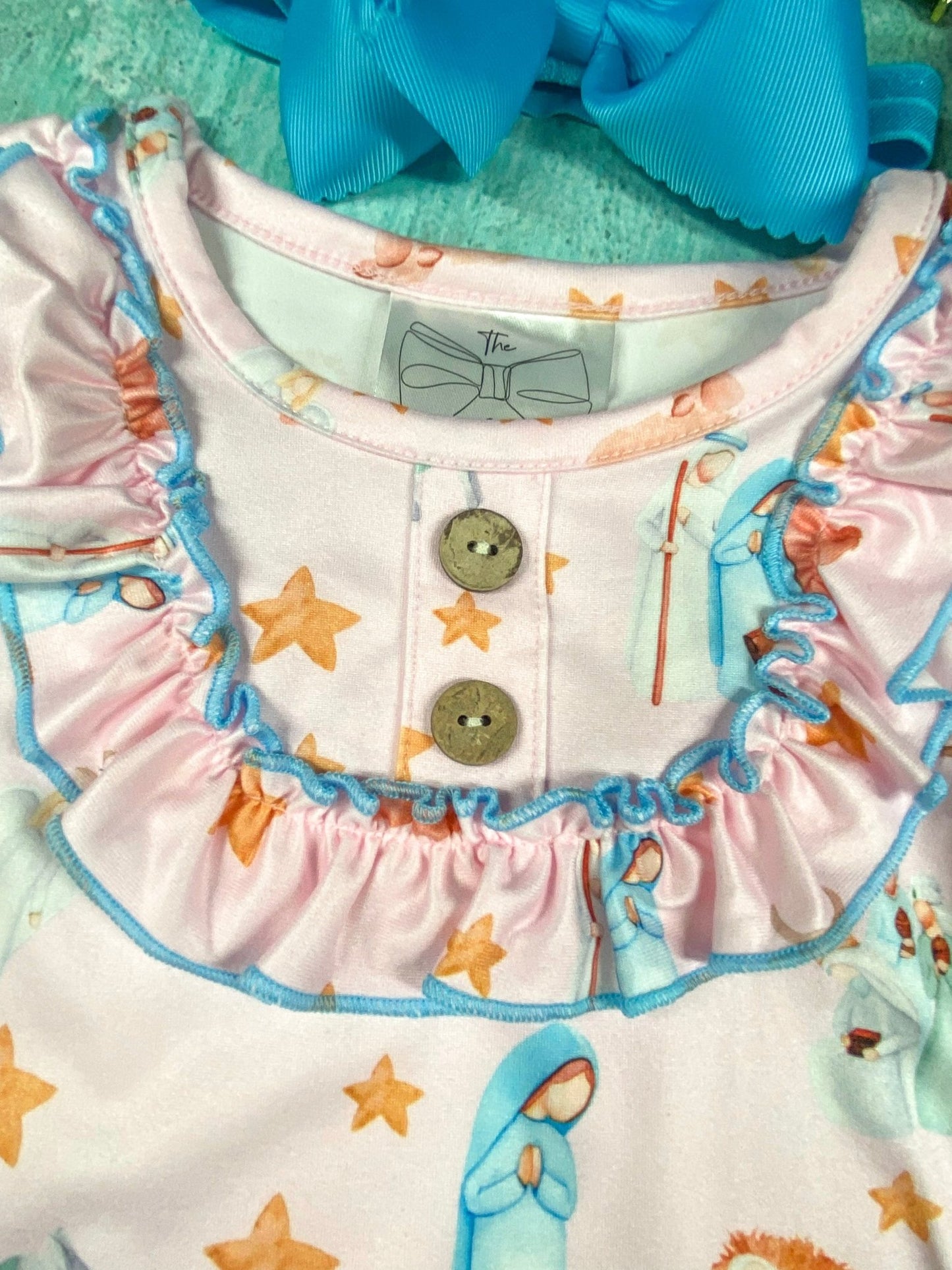 Close-up of a baby or toddler romper in light pink fabric with nativity-themed print featuring Mary, baby Jesus in a manger, stars, and animals. The neckline has ruffled trim outlined in blue stitching, two wooden buttons down the front, and a sewn-in clothing tag with a bow logo. A large blue bow accessory is partly visible above the garment.