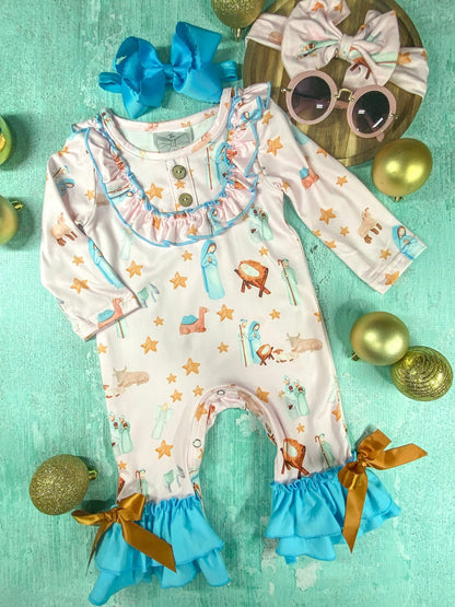 A long-sleeve baby romper with a light pink nativity print of Mary, baby Jesus, stars, and animals. The romper has ruffled trim around the neckline with two wooden buttons, blue ruffle cuffs with gold ribbon bows at the ankles, and a gathered waist. It is displayed on a turquoise background with Christmas ornaments, a large blue bow, and accessories including a matching headband and pink round sunglasses on a wooden board.
