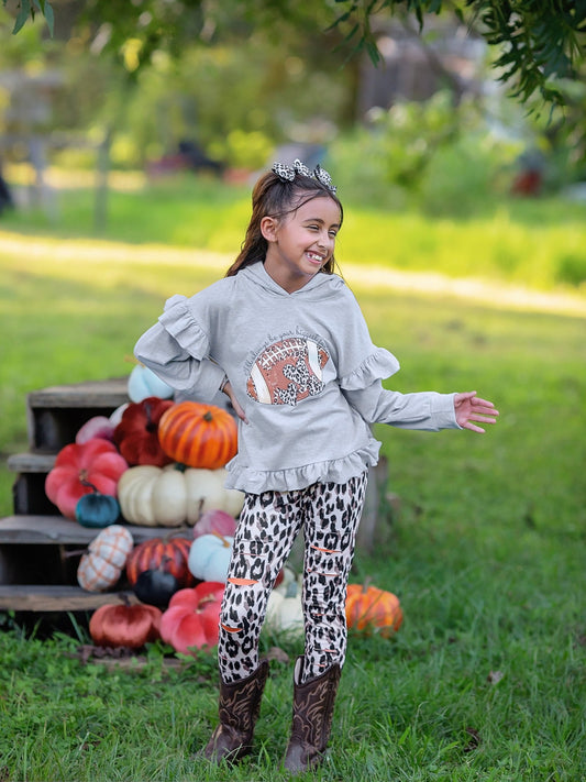 A child wearing a grey ruffled leggings set with a football pattern, standing in front of a display of pumpkins.
